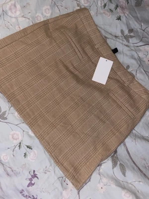 Beige rutig kortkjol - Säljer en beige kortkjol med rutigt mönster. Kjolen har bälteshällor och två dekorativa fickor baktill. Perfekt för dig som gillar klassisk stil med en modern twist.