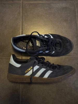Adidas Spezial svarta sneakers - Snygga Adidas Spezial sneakers i svart mocka med klassiska vita ränder på sidorna och guldfärgad text. Skorna har brun gummisula och svart snörning. Perfekt för dig som gillar retrostil och streetwear. Låg modell som passar till många outfits.