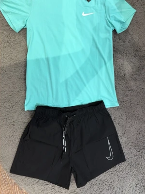 Nike träningsset turkos & svart - Snyggt träningsset från Nike med en turkos t-shirt i funktionsmaterial och svarta shorts med dragsko och Nike-logga. Perfekt för gymmet eller löprundan. T-shirten har rund hals och korta ärmar, shortsen har elastisk midja.