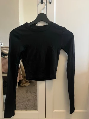 Svart croppad långärmad topp Gina Tricot - En svart croppad långärmad topp från Gina Tricot. Toppen har en enkel och stilren design med rund halsringning och är tillverkad i mjuk bomullsmix. Perfekt att matcha med höga jeans eller kjol.