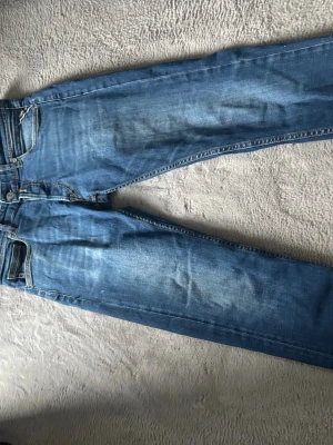 Blå jeans från Replay - Säljer ett par blå jeans från Replay med snygga slitningar och tvättad look. Klassisk femficksmodell med dragkedja och knapp i midjan. Jeansen har raka ben och är tillverkade i mjukt denimtyg som sitter skönt.