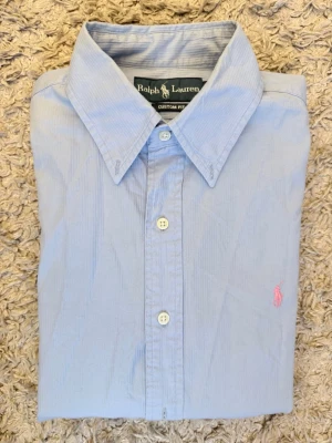 Ljusblå skjorta från Ralph Lauren - Klassisk ljusblå skjorta från Ralph Lauren med lång ärm och knappar framtill. Skjortan har en broderad rosa logga på bröstet och är tillverkad i bomull. Perfekt för en clean och stilren look.