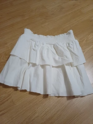 Vit volangkjol med resårmidja - Supersöt vit kortkjol med dubbla volanger och bred smockresår i midjan. Kjolen har en luftig och lätt känsla, perfekt för varma dagar. Passar till både sneakers och sandaler för en chill look.