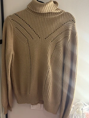 Beige stickad polotröja - Säljer en beige stickad polotröja med ribbad struktur och dekorativa hålmönster framtill och på ärmarna. Tröjan har lång ärm och hög polokrage, perfekt för lager på lager och en clean look. Passar dig som gillar stilrena och trendiga plagg. Står inget märke eller storlek men passar både S och M