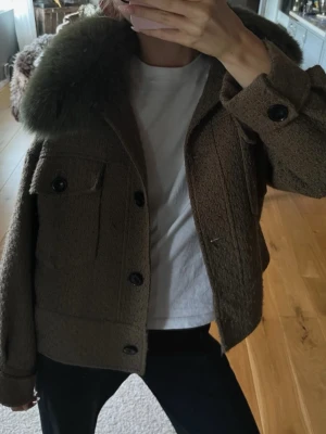 Meotine Olivgrön/brun pälsjacka - Säljer en olivgrön pälsjacka med stor fluffig krage. Jackan har en boxig passform, stora knappar framtill och två bröstfickor. Materialet är grovt och ger en cool vintagekänsla. Perfekt för kalla vinterdagar. I bra skick🙏🏼(mina bilder)