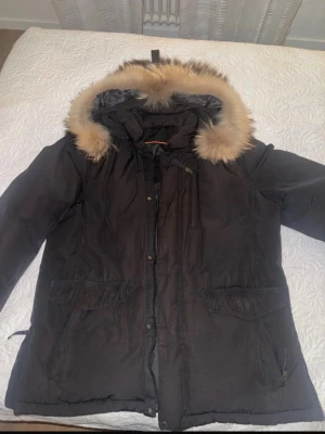 Svart  jacka med äkta päls - Svart parkas med stor huva och fluffig äkta päls i beige och bruna toner. Jackan har dragkedja och knappar framtill samt två stora fickor med lock. Perfekt för kalla dagar och riktigt mysig att bära.