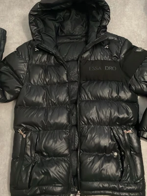 Svart pufferjacka Alessandro Zavetti - Helt perfekt skick, bara några bokstäver på bröstet borta. Snygg svart pufferjacka från Alessandro Zavetti med glansig finish, huva och dragkedjor i silver. Jackan har två framfickor med dragkedja och en patch med logga på ärmen. Perfekt för kalla dagar och riktigt trendig look.