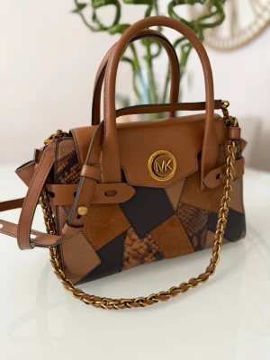 Patchwork handväska från Michael Kors - Snygg handväska från Michael Kors i patchwork-design med olika bruna och svarta nyanser. Väskan har detaljer i ormmönstrat och slätt läder, guldfärgade metalldetaljer och en kedjerem. 