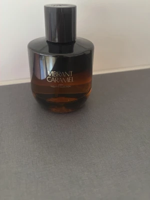 Vibrant Caramel parfymflaska - En elegant parfym från Zara med namnet Vibrant Caramel. Flaskan är rundad och har en snygg gradient från mörkbrunt till bärnstensfärgat glas. Locket är svart och flaskan har en modern, stilren design som känns lyxig och trendig.