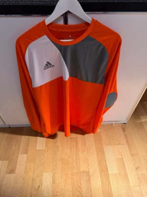 Orange långärmad tröja från Adidas - Snygg långärmad sporttröja från Adidas i orange med vita och grå detaljer. Tröjan har rund halsringning, gråa armbågslappar och Adidas-logga på bröstet. Tillverkad i ett lätt och funktionellt material, perfekt för träning eller match.