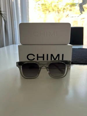 Grå CHIMI 04 - Snygga solglasögon från CHIMI med transparent grå båge och mörka glas. Rektangulär form som ger en modern vibe. Perfekta för soliga dagar och kommer med originaletui och box.