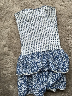 Blåvit axelbandslös kortklänning - Supersöt axelbandslös kortklänning i blått och vitt med smock upptill och volangkjol. Klänningen har ett unikt mönster med blå cirklar och linjer på vit botten. Perfekt för varma dagar och sommarens äventyr. Barn stolek!