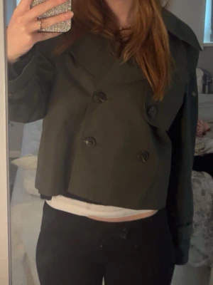 Kort mörkgrön trenchcoat - Säljer en kort trenchcoat i mörkgrönt tyg med dubbelknäppning och bred krage. Jackan har långa ärmar och en boxig, modern passform som slutar vid midjan. Perfekt för dig som vill ha en trendig och stilren look.