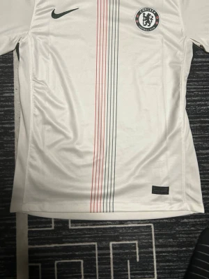 Chelsea vit matchtröja Nike - Chelsea FC:s vita matchtröja från Nike med tunna vertikala ränder i rött och svart på framsidan. Tröjan har klubbmärke på bröstet och svarta Nike-loggan. Tillverkad i lätt och ventilerande material, perfekt för fotbollsträning eller match.