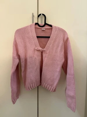 Rosa stickad kofta från Lindex - Söt ljusrosa stickad kofta från Lindex med V-ringning och knytband framtill. Koftan har långa ärmar och ribbstickade muddar vid ärmslut och nederkant. Perfekt för lager-på-lager och en gullig touch till din outfit.