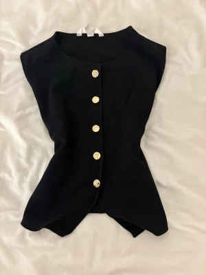 Svart ärmlös topp med guldknappar - Stilren svart ärmlös topp med rund halsringning och fem dekorativa guldknappar framtill. Toppen har en figurnära passform och är perfekt att styla till jeans eller kjol. Materialet känns mjukt och stretchigt.