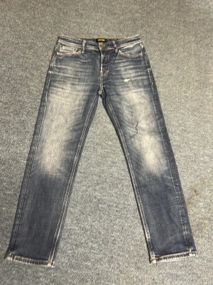 Blå tapered jeans från Jack & Jones - Snygga blå jeans från Jack & Jones i modellen Tapered/Mike. Jeansen har en tvättad look med slitningar och ljusa partier framtill. Klassisk femficksmodell och normalhög midja. Perfekta för en avslappnad stil.