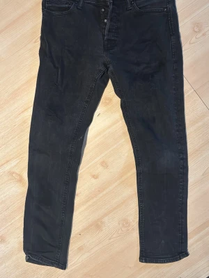 Svarta slim fit jeans - Säljer ett par svarta slim fit jeans med klassisk femficksdesign och knappgylf. Jeansen har diskreta slitningar på låret för en cool, avslappnad look. Tillverkade i mjuk denim med lite stretch för extra komfort.