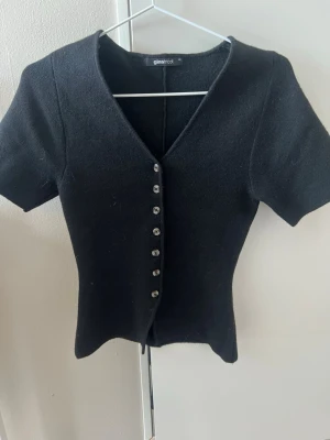 Svart kortärmad kofta från Gina Tricot - Stilren svart kofta från Gina Tricot med korta ärmar och djup V-ringning. Koftan har åtta knappar framtill och är gjord i ett mjukt, stickat material. Perfekt att matcha med jeans eller kjol för en trendig look.