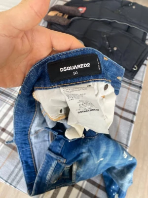 Blå Dsquared2 jeans med slitningar - Säljer ett par blå jeans från Dsquared2 i storlek 50. Jeansen har en cool tvätt med slitningar, färgstänk och trasiga detaljer för en trendig och avslappnad look. Klassisk femficksmodell med Dsquared2-logga bak i midjan. Perfekta för dig som gillar streetwear.