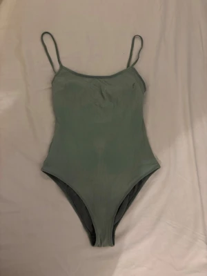 Grön ribbad baddräkt med smala axelband - Snygg grön baddräkt med ribbad struktur och smala axelband. Klassisk modell med rundad halsringning och hög benskärning. Perfekt för stranden eller poolen. Materialet känns stretchigt och bekvämt. 