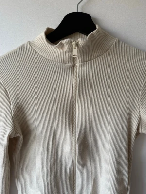 Beige ribbad ziptröja med hög krage - Säljer en beige ribbad tröja med dragkedja framtill och hög krage. Tröjan har lång ärm och är croppad i modellen, perfekt för en clean och stilren look. Materialet är stretchigt och mjukt, vilket gör den superbekväm att bära.