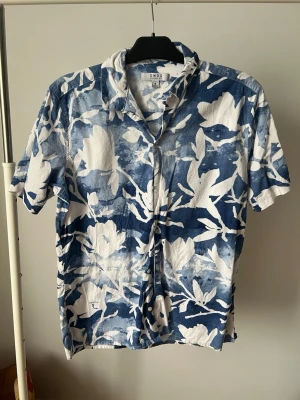 Blåvit blommig skjorta med kort ärm - Säljer en blå och vit skjorta med blommigt mönster och korta ärmar. Skjortan har klassisk krage och knappar framtill. Perfekt för dig som gillar en avslappnad och cool stil med lite extra färg. Materialet känns lätt och luftigt.