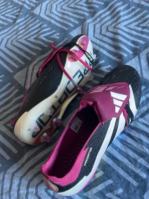Adidas Predator fotbollsskor svart/rosa - Snygga Adidas Predator fotbollsskor i svart, vitt och rosa med coola detaljer och text på sulan. Skorna har snörning, låg profil och greppiga dobbar för bästa fäste på planen. Ovandelen är i syntetmaterial och designen är modern och sportig.