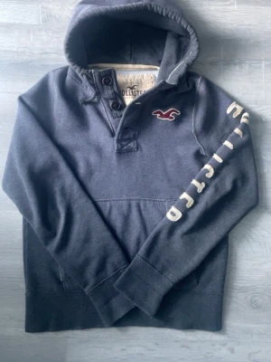 Hollister hoddie med knappar - Riktigt snygg och sällsynt hoddie från Hollister. Tröjan är väldigt eftertraktad och är svår att få tag på, knappar upp till och Hollister text på vänster arm gör verkligen tröjan unik från många andra! 