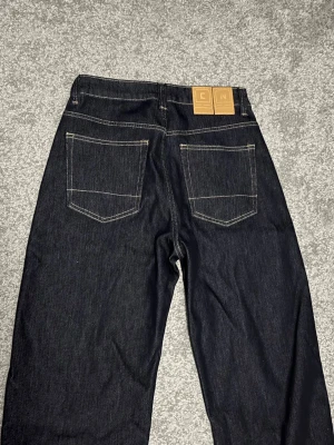 Mörkblå salvage Japan jeans  - Snygga mörkblå jeans från C.N med raka ben och klassisk femficksdesign, i storlek 30x32 baggy/rak fit. Helt nyskick! Och jeansen har kontrastsömmar och patch med logga bak i midjan. Tillverkade i kraftig denim som ger en stilren look.