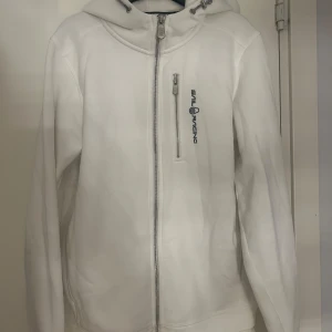 Vit zip hoddie från Sail Racing - Vit zip hoddie från Sail Racing med dragkedja framtill och en extra zipficka på bröstet. Jackan har en stilren look med logotyp på bröstet och justerbar huva med snörning. Perfekt för dig som gillar sportig och clean stil.