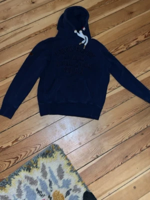 Mörkblå hoodie från Superdry - Mörkblå hoodie från Superdry med broderad text på bröstet och stora vita dragsnören i huvan. Klassisk känguruficka framtill och ribbade muddar vid ärmslut och nederkant. Perfekt för en avslappnad streetstyle.