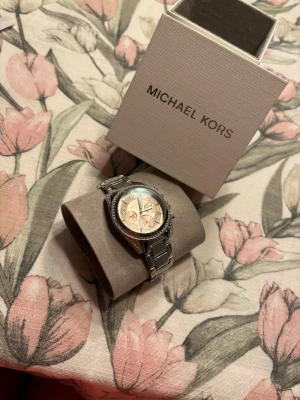 Silverfärgad klocka från Michael Kors - Stilren klocka från Michael Kors i silverfärg med länkarmband i metall. Urtavlan är ljus med flera små urtavlor och markerade siffror. Runt boetten sitter gnistrande stenar som ger extra lyxig känsla. Perfekt accessoar för att lyfta din outfit.