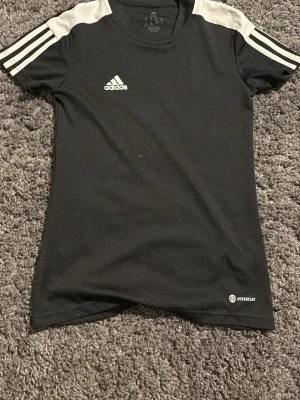 Svart Adidas träningströja barn - Snygg svart träningströja från Adidas med vita detaljer på axlarna och klassiska Adidas-ränder på ärmarna. Tillverkad i lätt och snabbtorkande material med AEROREADY-teknologi. Perfekt för träning eller sportaktiviteter. Pris kan diskuteras, barnstorlek!