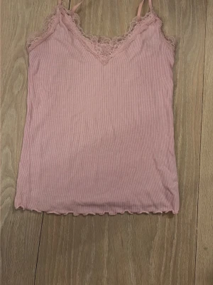 Rosa ribbad spetslinne - Säljer ett ljusrosa ribbat linne med tunna axelband och vacker spetsdetalj längs v-ringningen. Materialet är mjukt och stretchigt, perfekt för lager på lager eller sommarens outfits. Linnet har vågig kant nertill för en extra söt touch.