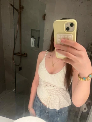 Vit somrig ribbad linnetopp med spetsdetaljer - Söt vit linnetopp med ribbad struktur, spetsdetaljer och volang nedtill. Toppen har smala axelband med rosset knyt och v-ringad hals. Perfekt för sommarens alla tillfällen och ger en romantisk vibe. Samt stand vibe 