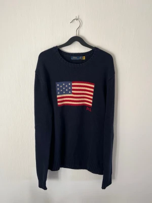 Mörkblå stickad tröja Polo Ralph Lauren USA flagga - Mörkblå stickad tröja från Polo Ralph Lauren med amerikansk flagga i rött, vitt och blått på bröstet samt RL-broderi. Tröjan har rund halsringning, och ribbade muddar. Storlek Large