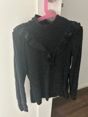 Glittrig svart topp med volang Lindex - Snygg svart topp från Lindex med glittriga detaljer och volang framtill. Hög hals och långärmad modell som ger en trendig look. Perfekt för dig som vill sticka ut lite extra med en touch av glitter.