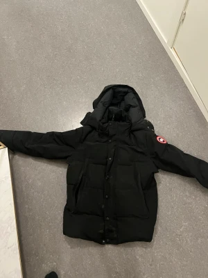 Svart dunjacka från Canada Goose - Säljer en svart dunjacka från Canada Goose med huva och stora framfickor. Jackan har det klassiska runda Canada Goose-märket på ärmen och stängs med både dragkedja och knappar. Perfekt för kalla vinterdagar och riktigt varm.