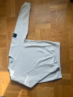 Beige sweatshirt från Stone Island - Snygg beige sweatshirt från Stone Island med klassisk rund halsringning och ribbade muddar. Tröjan har en enkel och stilren design, perfekt för dig som gillar minimalistiskt mode. Tillverkad i mjukt bomullsmaterial för skön komfort.