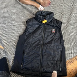 Svart dunväst från Parajumpers - Snygg svart dunväst från Parajumpers med hög krage och gul detalj vid dragkedjan. Västen har två framfickor med dragkedja och ett broderat Parajumpers-märke på bröstet. Perfekt för lager-på-lager och streetwear-stil.