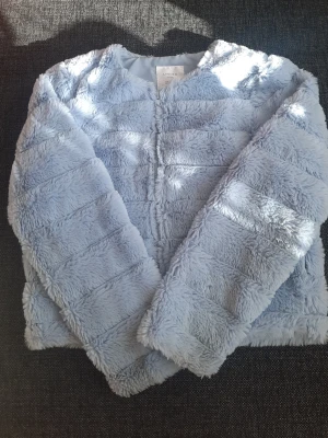 Ljusblå pälsjacka från Lindex Kids - Supermjuk ljusblå pälsjacka från Lindex Kids i storlek 146/152. Jackan har fluffig fuskpäls, rund halsringning och stängs framtill. Perfekt för att hålla sig varm och snygg under kalla dagar.