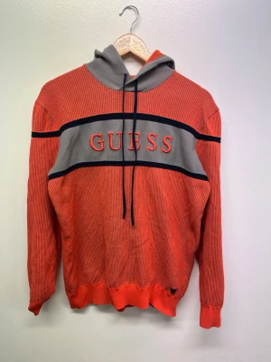 Röd och grå hoodie från Guess - Snygg röd hoodie från Guess med grå huva och bred grå panel över bröstet där det står GUESS i stora bokstäver. Svarta detaljer och ribbade muddar vid ärmar och nederkant. Tillverkad i ett mjukt, ribbat material som passar perfekt för kalla dagar.