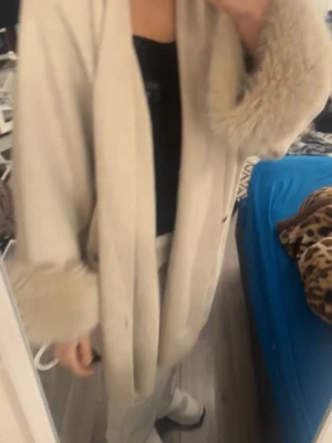 Beige lång rock med fuskpälsdetaljer - Stilren beige lång cardigan med breda slag och fluffiga fuskpälsdetaljer vid ärmslut och krage. Rak passform och öppen framtill, perfekt för en trendig och elegant look. Passar till lager-på-lager och ger en lyxig känsla. One size- passar S-M