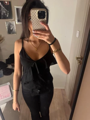 Svart linnetopp med volangdetalj - Snygg svart linnetopp med tunna axelband och v-ringning. Toppen har en volangdetalj framtill som ger en extra touch. Perfekt att matcha med jeans eller kjol för en trendig look.