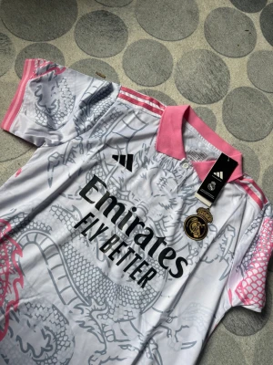 Real Madrid Adidas matchtröja rosa/vit - Säljer en Real Madrid matchtröja från Adidas i vitt med rosa detaljer och drakmönster. Tröjan har krage, knappar och klubbmärke på bröstet samt svarta logotyper och sponsortryck. Slim fit-modell i funktionsmaterial.