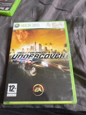 Need for Speed Undercover Xbox 360 - Säljer spelet Need for Speed Undercover till Xbox 360. Omslaget har en cool stadssilhuett i orange och svart med snabba bilar och palmer. Perfekt för dig som gillar racing och action. PEGI 12+. Fodralet är grönt och spelet är från EA.
