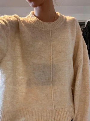 Beige stickad tröja med ribbad kant - Mysig beige stickad tröja med rund halsringning och ribbade muddar vid ärmslut och nederkant. Tröjan har en dekorativ söm framtill och är lätt oversized med långa ärmar. Perfekt för lager på lager och chill dagar.