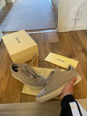 Gråa Axel Arigato sneakers med dragkedja - Snygga gråa sneakers från Axel Arigato i mocka med unik dragkedja framtill istället för klassiska snören. Skorna har en låg profil och vit sula som ger en clean look. Perfekta för dig som gillar stilrena och moderna detaljer.