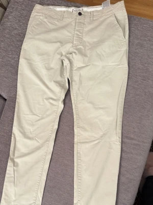 Beige chinos från Jack & Jones - Snygga beige chinos från Jack & Jones med klassisk passform och raka ben. Byxorna har fickor fram och bak samt stängs med knapp och dragkedja. Perfekta för en stilren och avslappnad look.